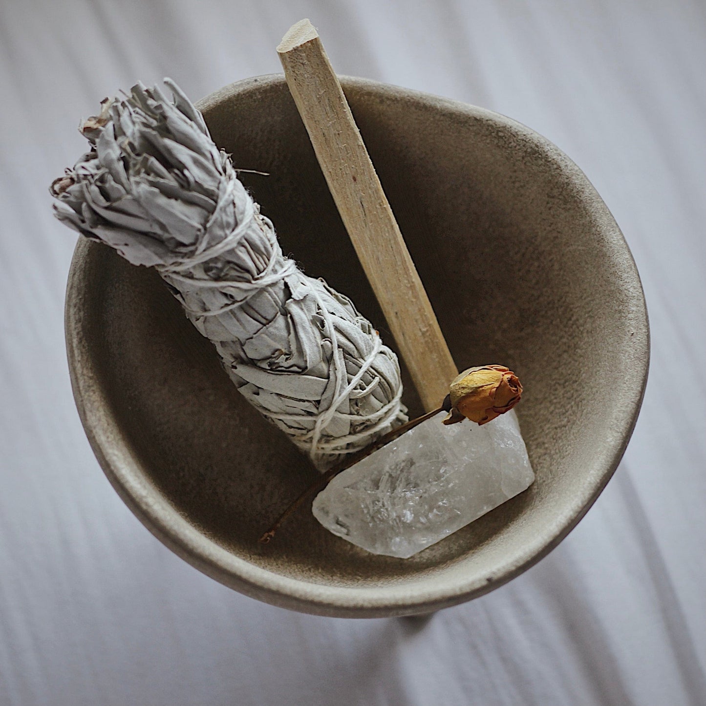 A photo showing a light brown ceramic bowl, contains a white sage sticks, a crystal, a dried flower bud, to use in the ritual of smudging.